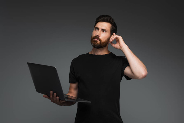 pensive freelancer with beard holding laptop and looking away isolated on grey - Foto, Imagen