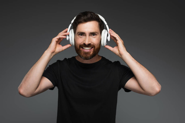 cheerful and bearded man in wireless headphones listening music isolated on grey - Фото, изображение