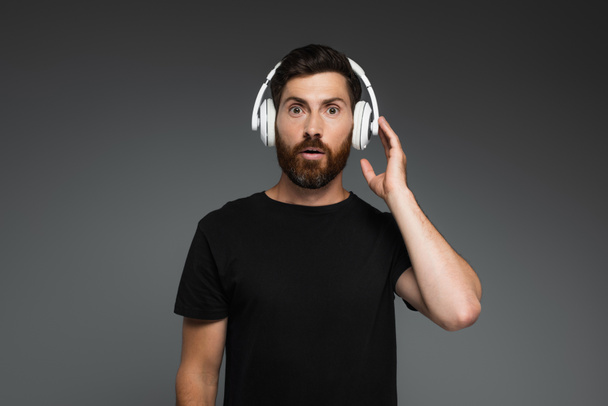 surprised man touching wireless headphones and listening music isolated on grey - Fotó, kép