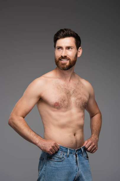 joyful and shirtless man with beard adjusting denim jeans isolated on grey - Fotó, kép