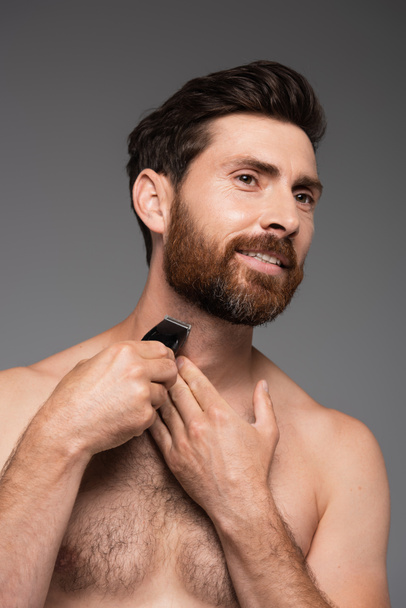 portrait of bearded and shirtless man with hair on chest shaving with electric razor isolated on grey - Foto, Imagem