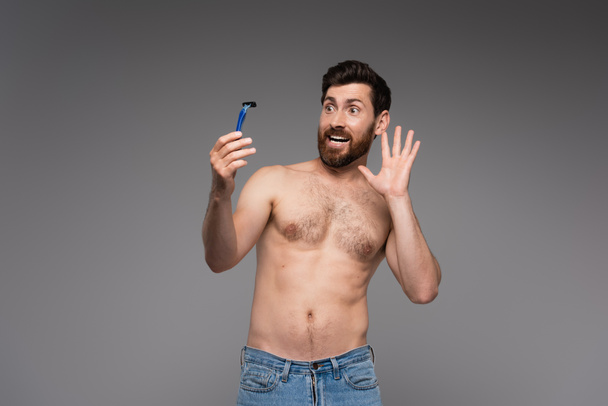 испуганный и без рубашки человек с бородой держа безопасную бритву изолированы на серый - Фото, изображение