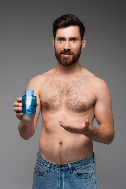 bearded and shirtless man pointing with hand at bottle with after shave product isolated on grey - Fotoğraf, Görsel