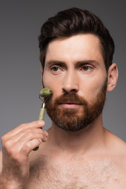 портрет человека без рубашки с бородой с помощью нефритового ролика во время массажа лицо изолированы на сером - Фото, изображение