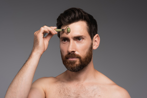 shirtless man with beard using jade roller while massaging forehead isolated on grey - Fotó, kép