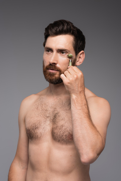 Мужчина без рубашки с бородой с помощью нефритового роллера во время массажа лицо изолированы на сером - Фото, изображение