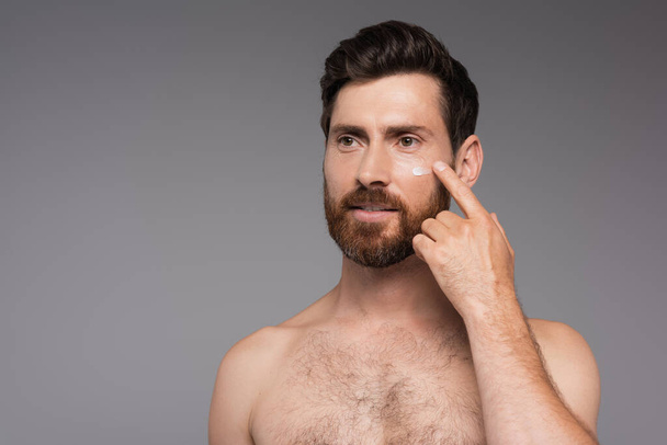 bearded and shirtless man applying cream on face isolated on grey - Foto, imagen