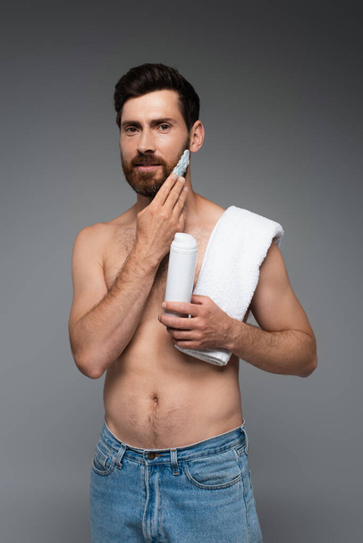 бородатый человек с полотенцем применяя пену для бритья и глядя на камеру изолированы на сером - Фото, изображение