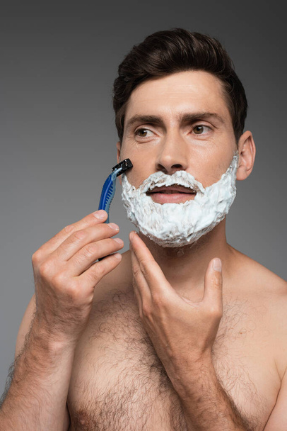 shirtless man with white shaving foam on face shaving with safety razor on grey - Photo, image