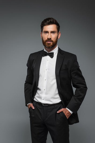 bearded man in elegant tuxedo with bow tie posing with hands in pockets isolated on grey  - Foto, Imagen
