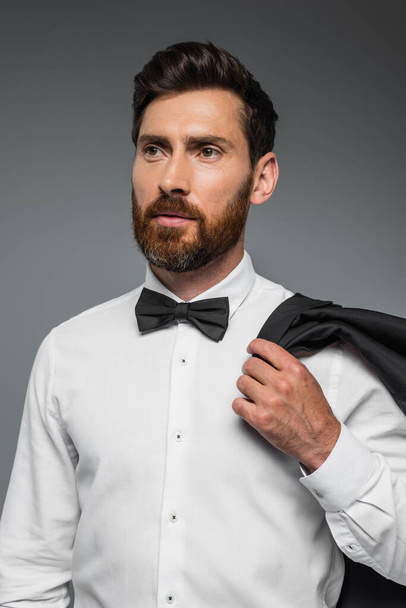 portrait of bearded man in suit with bow tie holding blazer isolated on grey  - Fotoğraf, Görsel