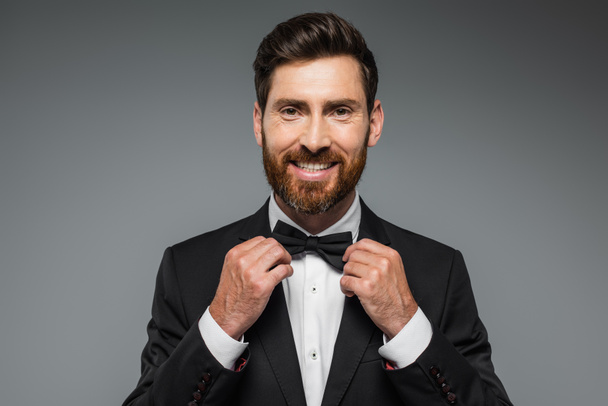 bearded man in suit smiling and adjusting bow tie isolated on grey  - Foto, imagen