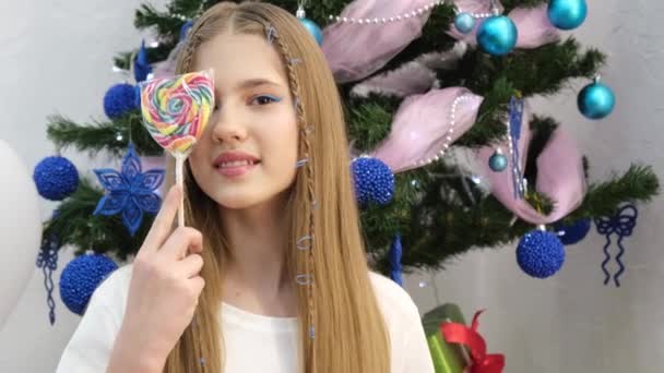 Happy teen girl in white sweater with lollipop tsandi on a stick on the background of the Christmas tree, caramel shop. - Metraje, vídeo