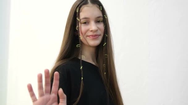 European teen girl waves her hand at the camera, she is on a white background. Beautiful child of 12 years smiling, long straight hair - Materiał filmowy, wideo
