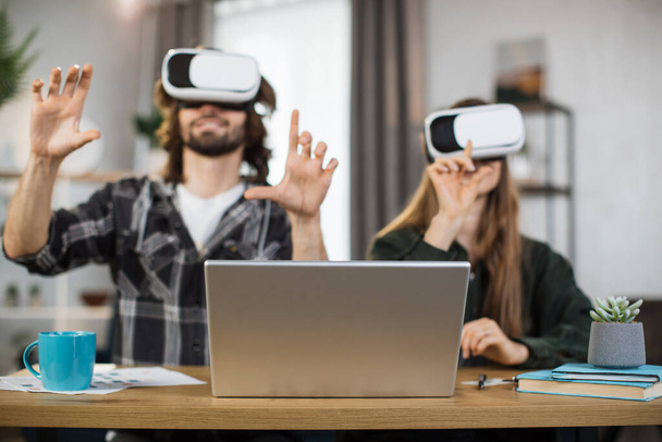 Excited young people with surprised expression, playing games at home, using VR glasses and laptops. Happy family enjoying artificial reality entertaining themselves with innovations. - 写真・画像
