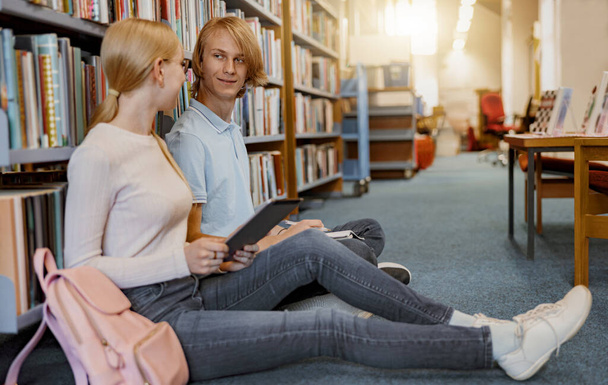 Friends student sit on floor near bookshelves in library and studying. Education concept - 写真・画像