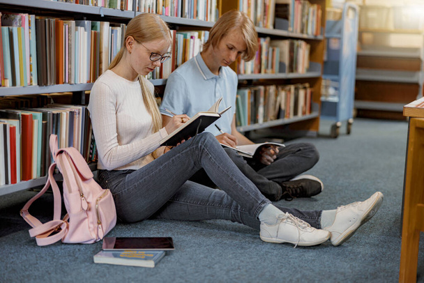 Друзі студенти сидять на підлозі біля книжкових полиць у бібліотеці та навчаються. Концепція освіти
 - Фото, зображення