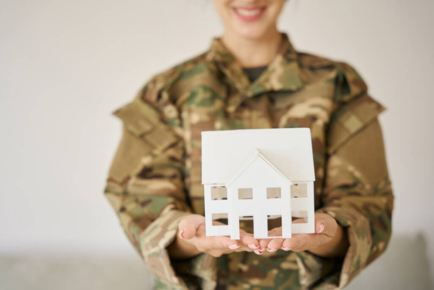 White toy homemade house in the hands of a female soldier in camouflage uniform on a light background - Foto, Bild