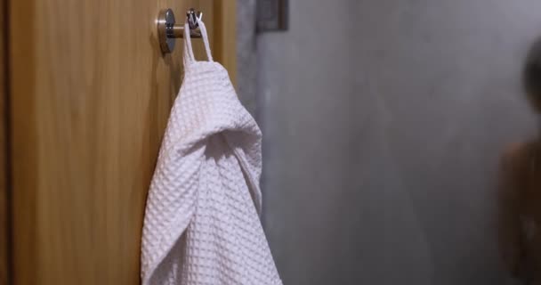 White robe on hook and naked woman taking relaxing shower in morning or evening. Concept of body hygiene and services of spa centers and hotels - Imágenes, Vídeo