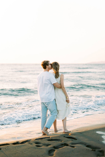 Man hugs and kisses woman on the temple while standing on the beach. Back view. High quality photo - 写真・画像