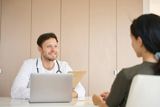 Optimistic doctor communicates kindly with a young patient in a bright office, tablet and a laptop are on the table - 写真・画像
