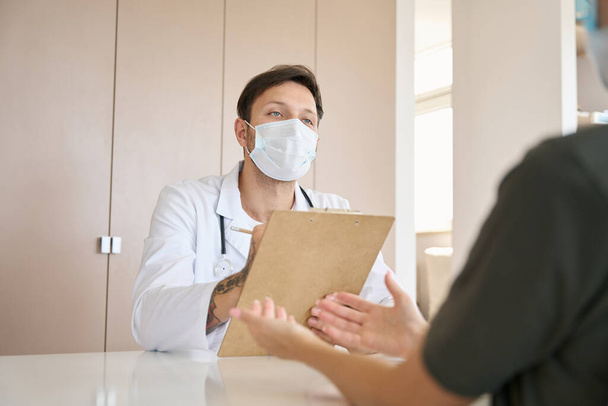 Γιατρός με λευκό παλτό και προστατευτική μάσκα ακούει προσεκτικά και καταγράφει τα παράπονα των ασθενών γραπτώς. - Φωτογραφία, εικόνα