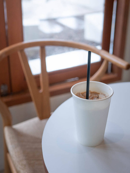 Кофе со льдом в белой простой пустой бумажной чашке на столе возле окна в минимальном японском стиле кафе - Фото, изображение