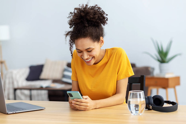 Young African American woman using mobile phone, chatting online sitting at a desk in a home office. Female freelancer texting online using smartphone and smiles friendly - Foto, Imagem