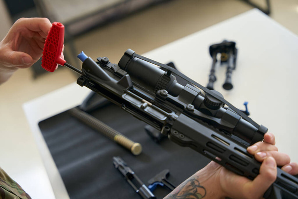 Ο άνθρωπος καθαρίζει ένα αποσυναρμολογημένο πυροβόλο όπλο με ένα ειδικό εργαλείο σε μια χαρτοπετσέτα, μέρη του πολυβόλου που ορίζονται - Φωτογραφία, εικόνα