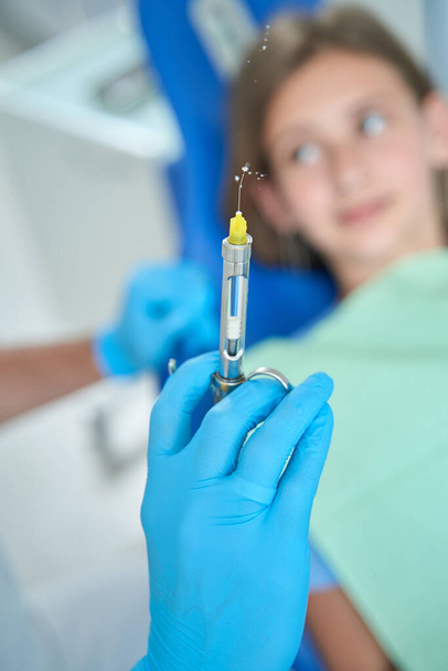 Adolescent girl lying in dental chair and looking at pediatric dentist with syringe in hand - 写真・画像