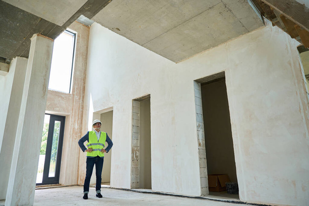 Adult male foreman stands in huge unfinished house with protective helmet on his head and wearing a green construction vest - Valokuva, kuva