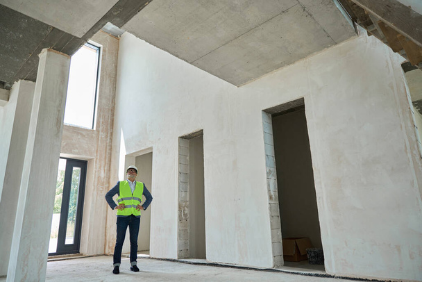 Serious male foreman inspecting a huge unfinished house with a safety helmet on his head and wearing a green construction vest - Foto, immagini