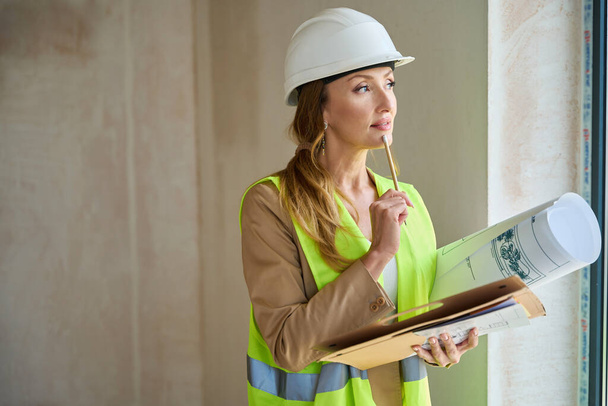 Thoughtful woman realtor holds a pencil near her face, and in her hands she holds packs and rolls of blueprints - Foto, Imagen