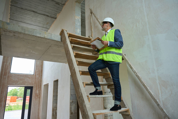 Мужчина-мастер в защитном шлеме и зеленом жилете поднимается по деревянной лестнице в недостроенном доме - Фото, изображение