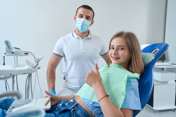Happy teenage girl seated in dental chair beside pedodontist making thumbs-up gesture - Fotó, kép