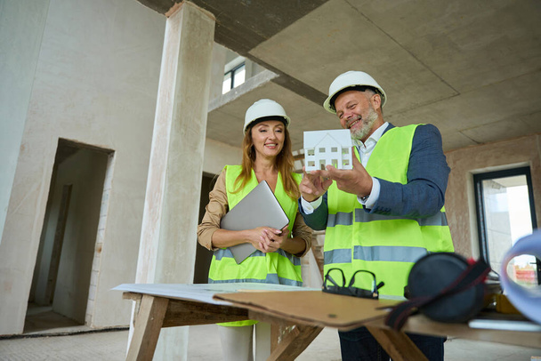Joyful foreman showing a woman realtor with a laptop in hand a miniature of a house under construction - Fotó, kép