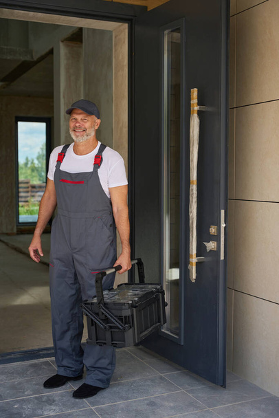 Portrait of an adult smiling builder in overalls and with a tool box. Man stands in front of the front door - Fotografie, Obrázek