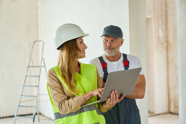 Agent communicates with adult worker hold open laptop in hands. Man and woman standing at a construction site - Φωτογραφία, εικόνα