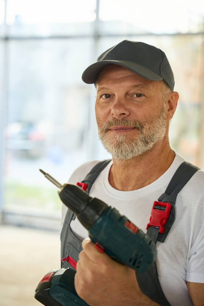 Portrait of a bearded builder with a drill in his hands. Man at a construction site - Foto, imagen
