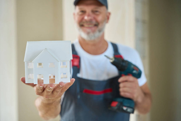 Photo of smiling construction worker in baseball cap and overalls holding house model in one hand and drill in other - Фото, изображение