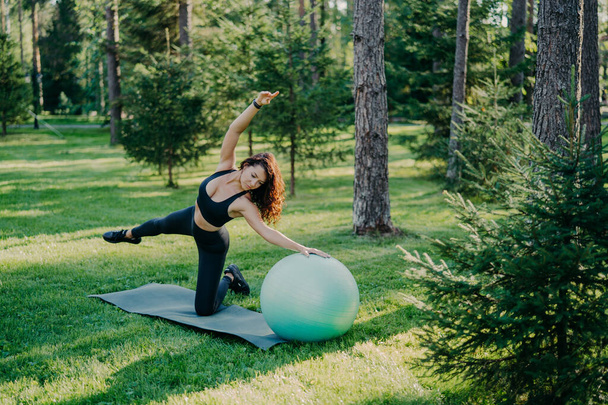 Fitness, sport and healthy lifestyle concept. Outdoor shot of slim brunette European woman does pilates and balance exercise with fit ball tilts aside poses in green forest on grass at karemat. - Photo, Image