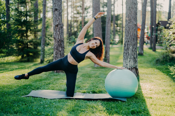 Horizontal shot of active slim woman with cheerful expression leans aside does exercises with fitness ball, dressed in sportsclothes, poses outdoor against tall trees on karemat. Healthy lifestyle - Fotó, kép