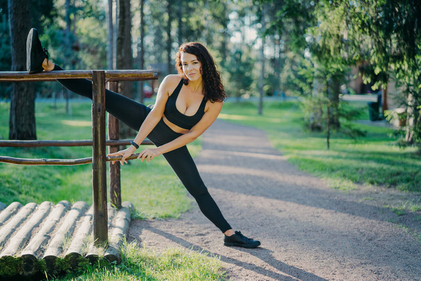Flexibele jonge krullende vrouw draagt zwarte top legging en sneakers, strekt benen, sport tijdens de zomerdag in de open lucht, poseert tegen groene bomen. Mensen, flexibiliteit en trainingsconcept - Foto, afbeelding