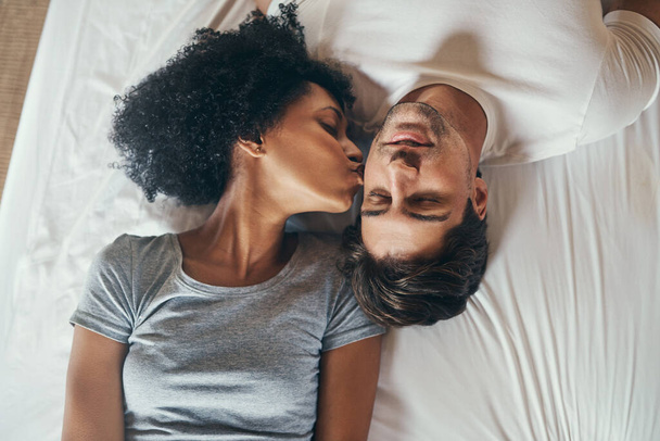 Wakey, wakey sleepy head. High angle shot of a young woman kissing her boyfriend on the cheek while lying in bed together at home - Photo, image