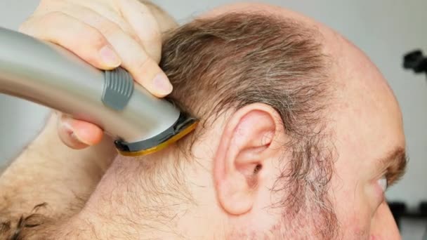 Man shaves his head. An overgrown man with a bald patch clumsily cuts his brown gray hair on his head with an electric clipper. Self shaving head with a trimmer. Amateur haircut in the bathroom - Imágenes, Vídeo