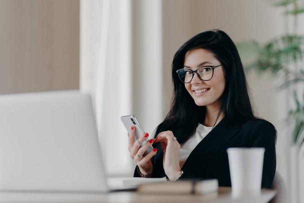 Horizontal shot of happy brunette European woman messages on modern smartphone, gives online consultation, smiles positively, wears spectacles and formal suit, sits in front of opened laptop. - Photo, Image