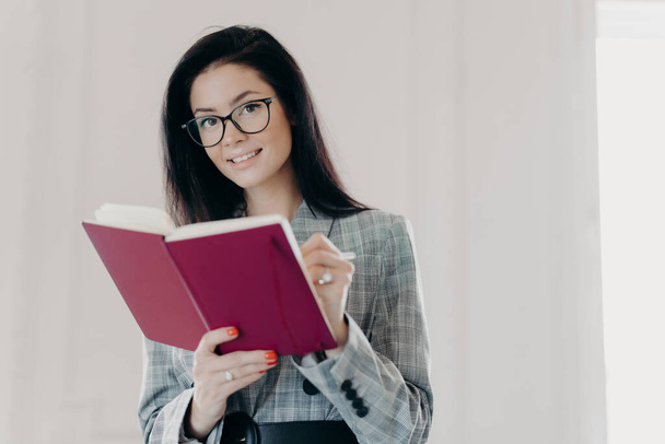 Beautiful business teacher plans her lessons, educates high school student, makes notes in notepad, wears spectacles and formal clothes, poses against white background. Woman writes down plans - Foto, Bild