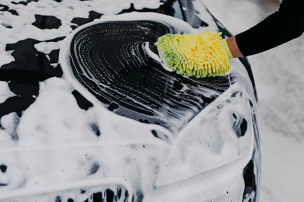 Mans mão com esfregão carro de limpeza com auto shampoo. Sabão suds em capô preto. Conceito de veículo. Automóvel moderno coberto por espuma - Foto, Imagem