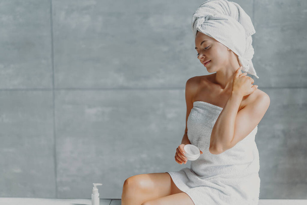Satisfied spa woman sits in profile applies body cream for perfect skin, enjoys pampering and beauty treatment poses wrapped in towel against grey wall in bathroom. Taking good care of own body - Photo, Image