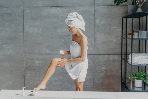Indoor shot of slim female model applies leg cream stands wrapped in bath towel takes care of body and skin undergoes beauty treatments after taking shower poses in bathroom. Cosmetology concept - Foto, immagini